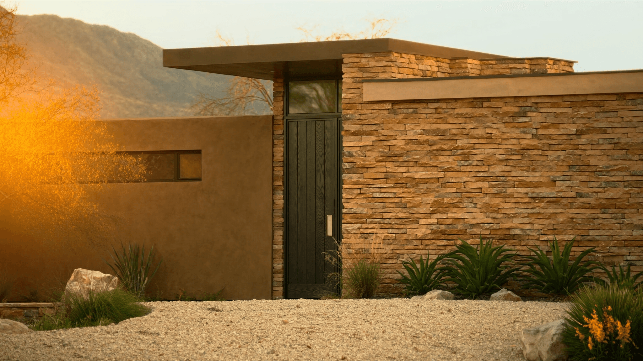 The stone-and-stucco entrance of a modern Southwest custom home at golden hour in El Paso, Texas, with a dark wood door, desert xeriscape, and the Franklin Mountains behind