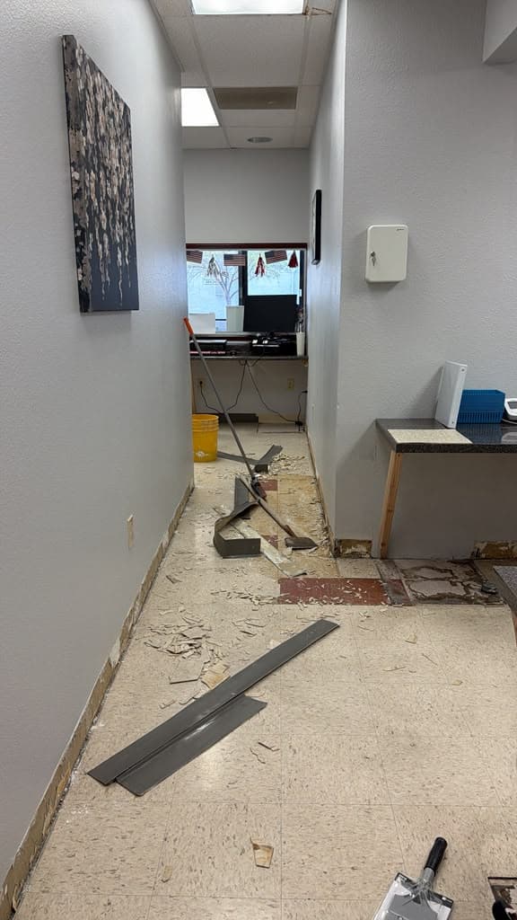 Existing vinyl composition tile being scraped up by hand in a medical office corridor, with debris and a floor scraper visible mid-demolition.