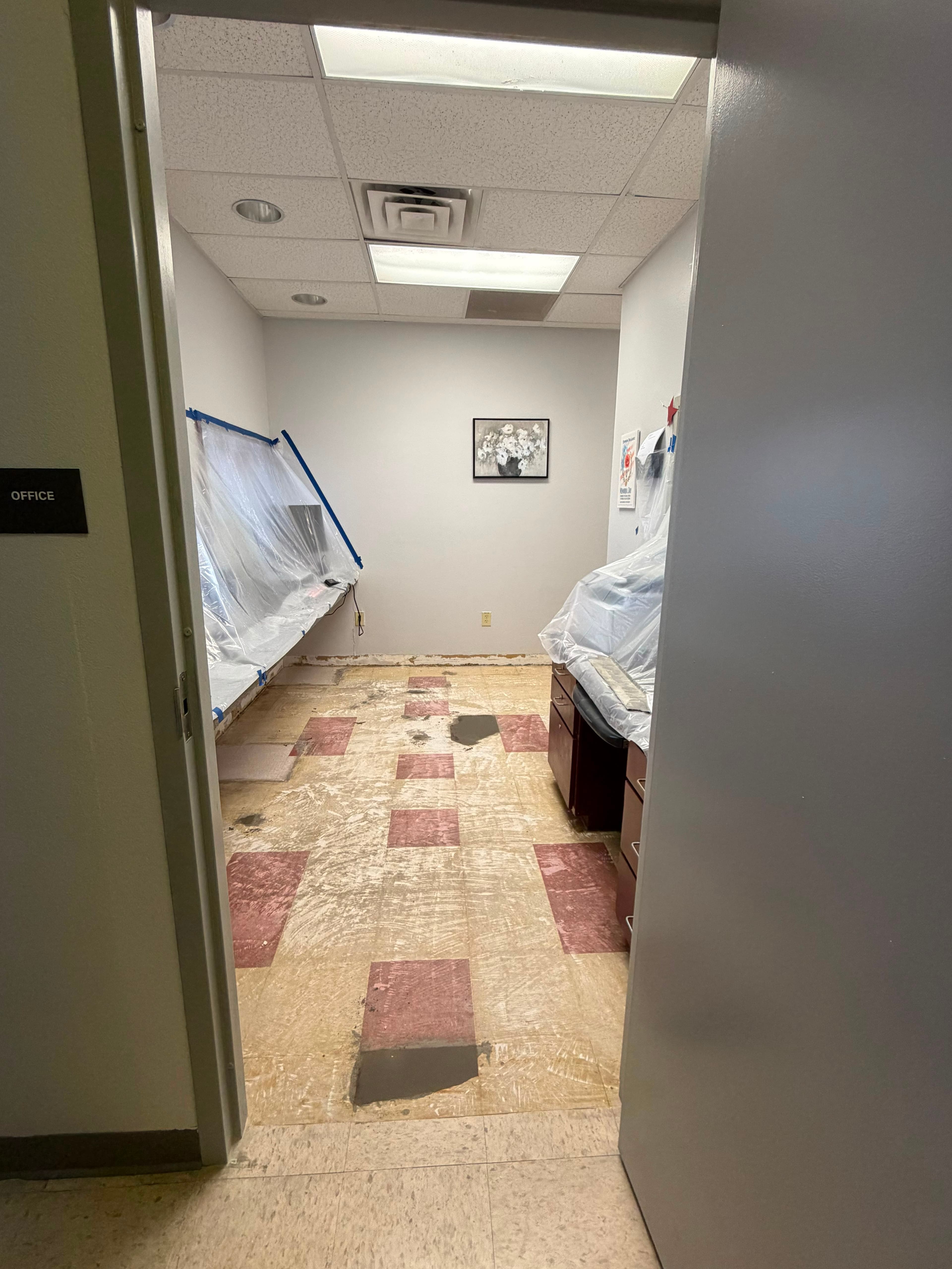An office with furniture and cabinetry wrapped in plastic and secured with tape, floor stripped to the underlayment, during a Sage Intelligence commercial flooring replacement.